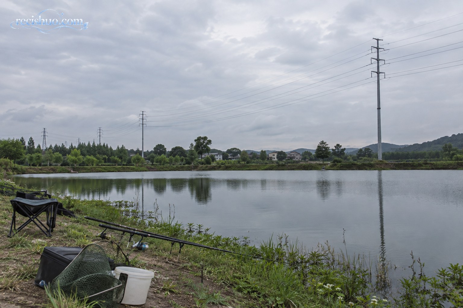 阴天乡村鱼塘岸边放着钓具,上方架空电力线路清晰可见