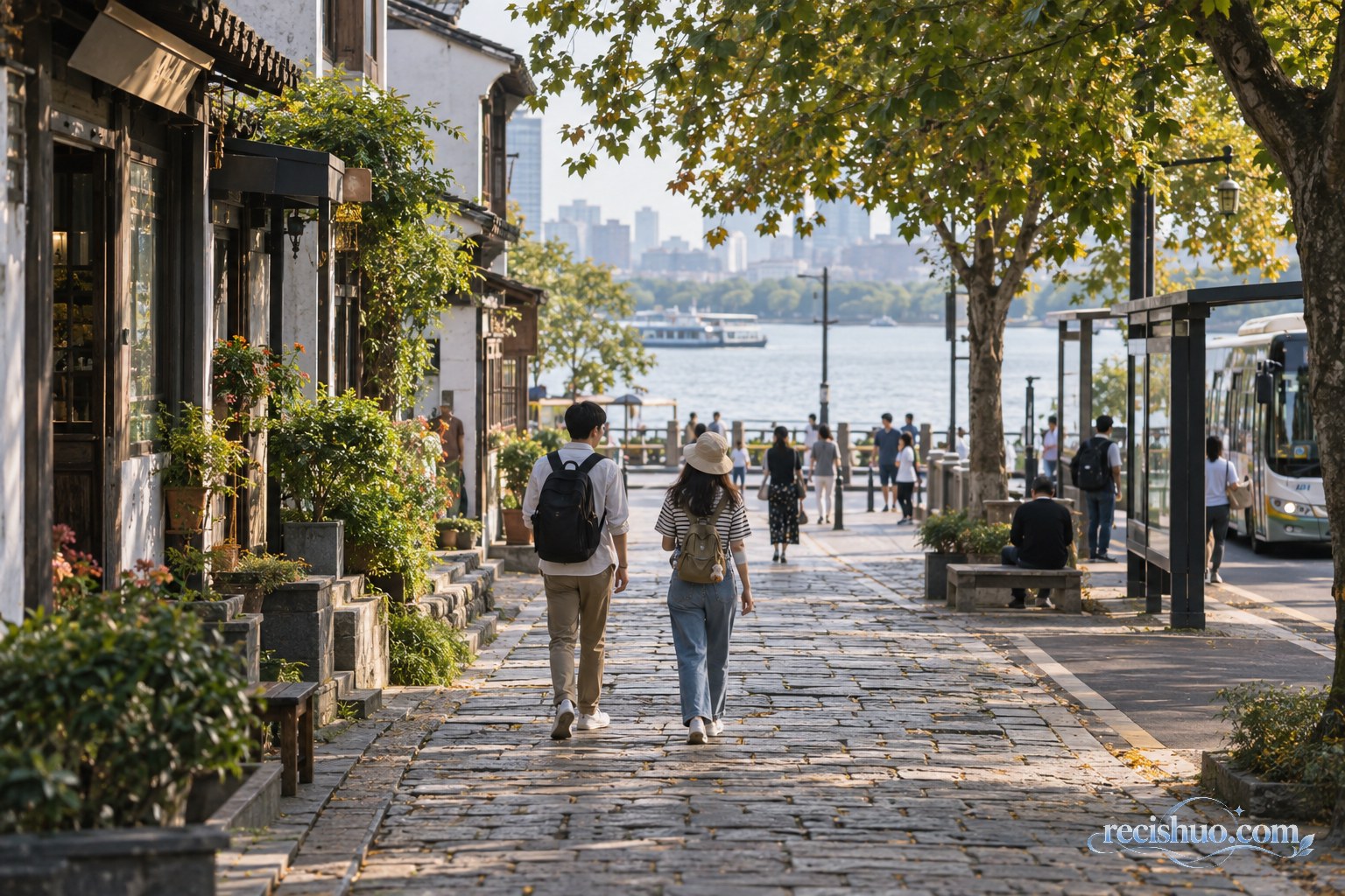 两名背着轻便小包的游客沿滨水老街步行,附近有树荫、公交站和日常生活场景