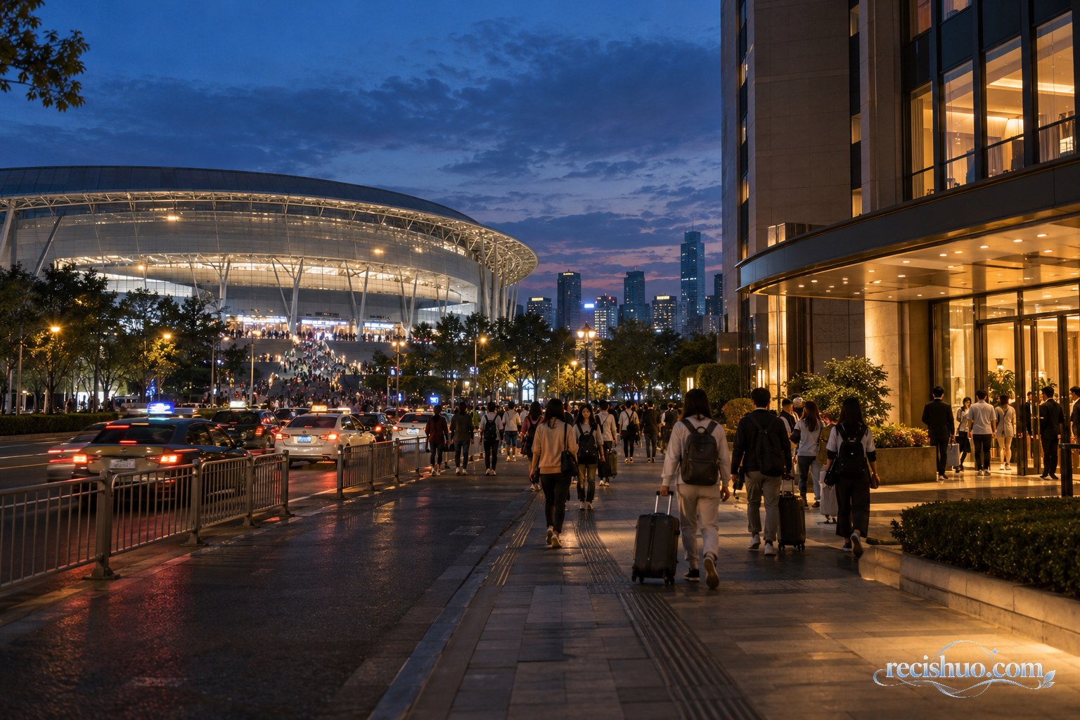 夜幕下体育馆迎来观众,邻近酒店亮灯并有旅客携行李经过